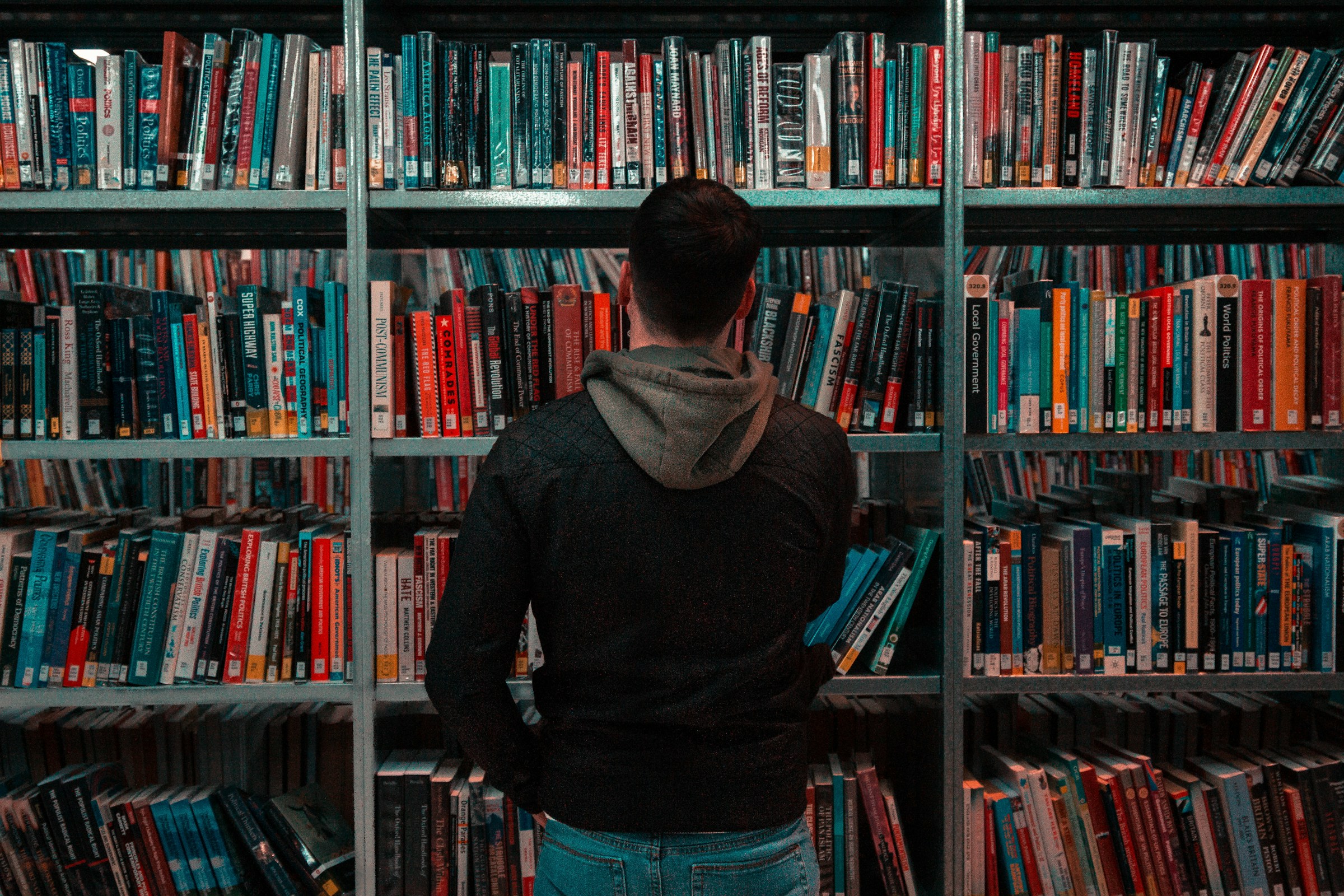 Imagen de portada para la columna de Jennifer Cabrera sobre los cambios al Fondecyt de Postdoctorado - En ella, se ve un gran estante lleno de libros, con una persona en el frente y dando la espalda a la cámara, buscando un libro en particular. La imagen representa el conocimiento general.