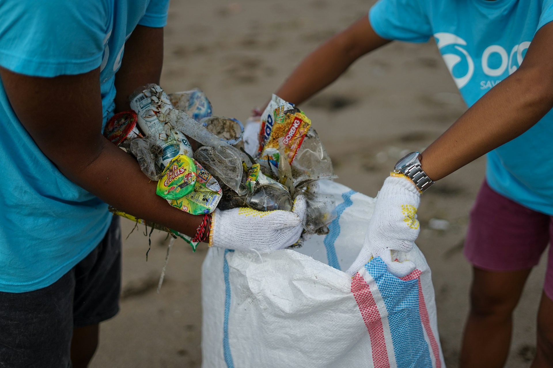 La imagen muestra a dos personas depositando residuos de una playa en una bolsa. Imagen de OCG Saving the ocean